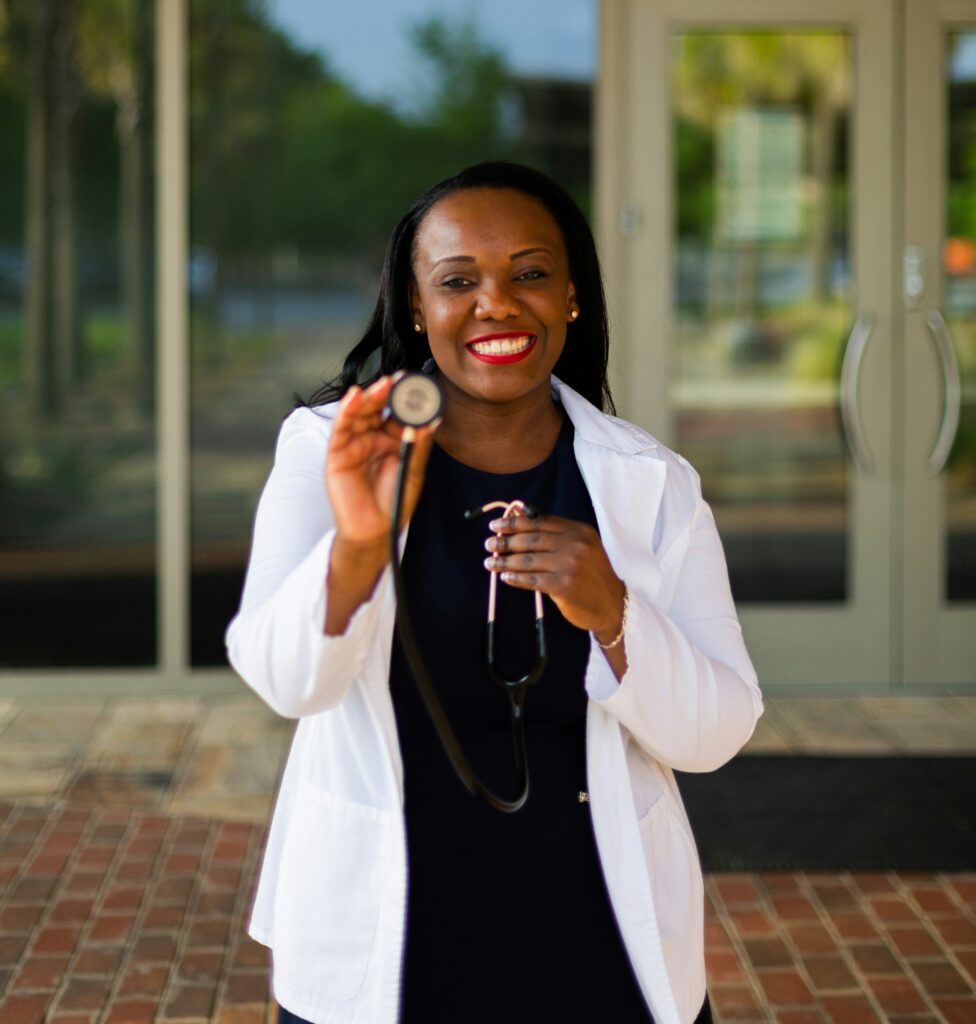 Smiling healthcare professional in a white coat holding a stethoscope outside a medical office.